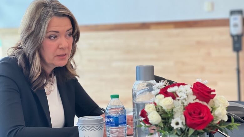 Woman seated at table with a microphone, flowers, water bottle and portable coffee cup in front of her.