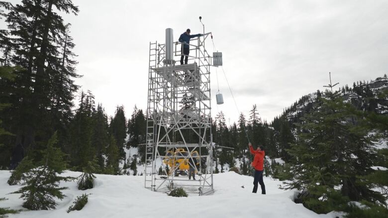 Two people work on a large metal tower with a helicopter and snowy mountains in the background.