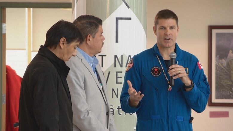 Two Indigenous men from Turtle Lodge stand next to astronaut Jeremy Hansen, who is holding a microphone.