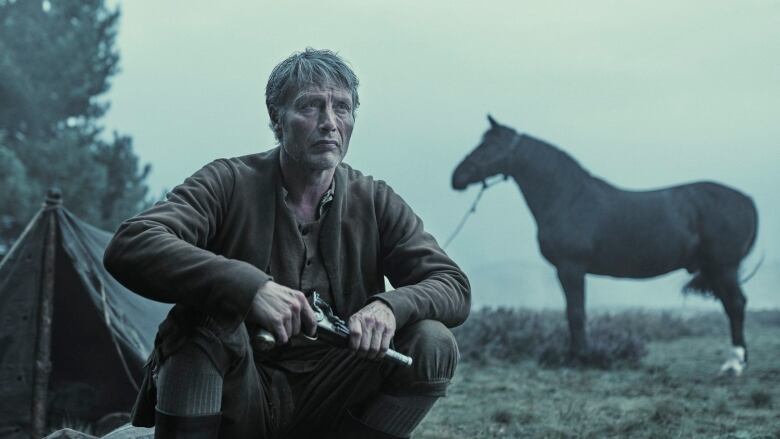 A man holding a pistol squats in front of a horse and a tied horse. Behind him is a foggy field filled with shrubs.