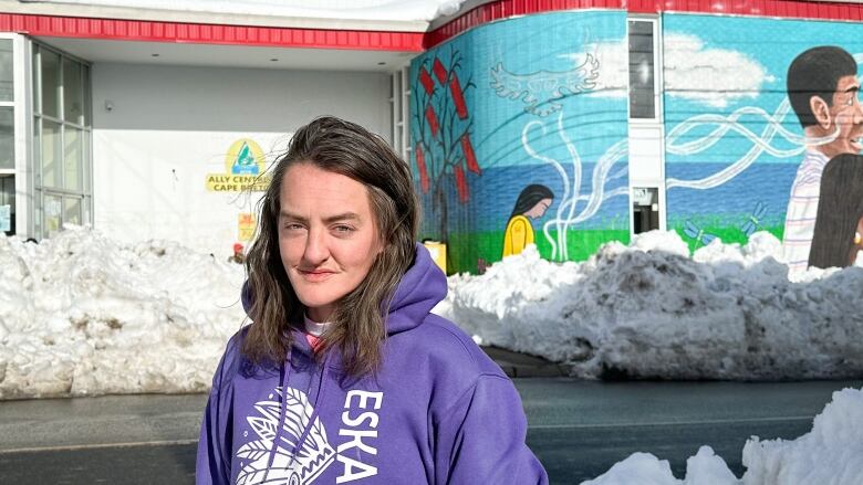 A woman in a purple hoodie with Eskasoni on it stands in front of a building with a colourful mural on it and blocked by snow piles.