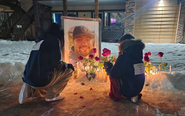 Two people kneel down in front of a framed photo of a man's face.