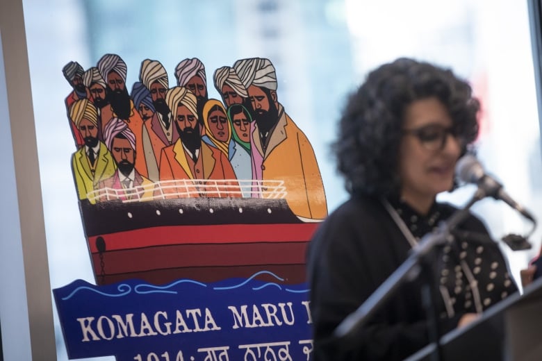 A South Asian woman speaks in front of an artwork depicting a number of South Asian people looking sad above a ship's hull.