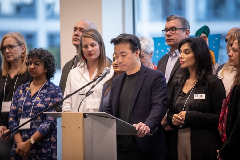 An East Asian man stands at a podium while being flanked by a number of others.