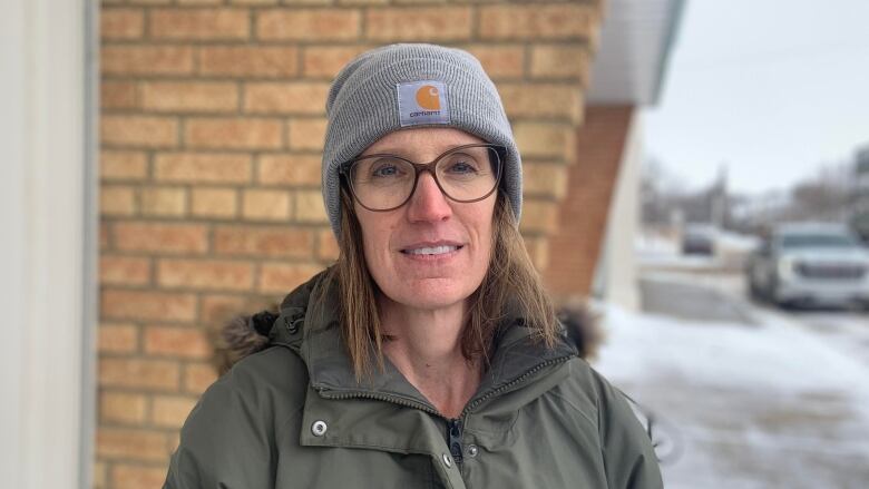 A woman in a sage winter coat and grey Carhartt toque stands outside on a sidewalk in wintertime. 