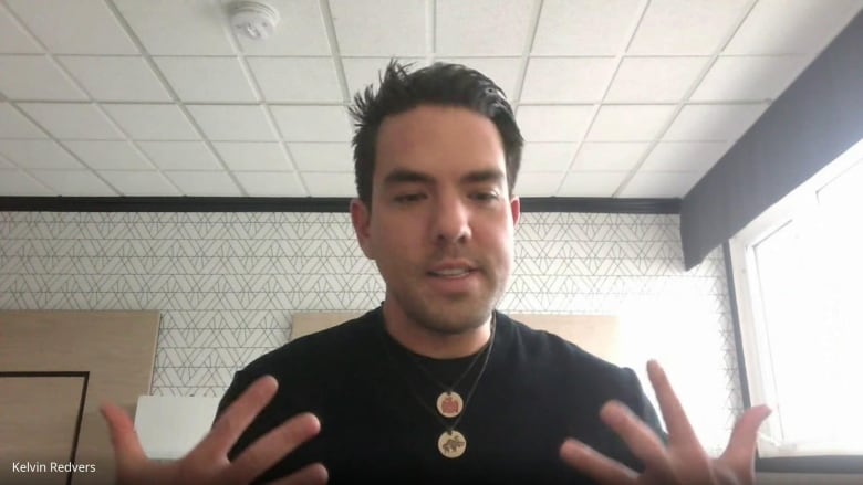 A man sits in a hotel room. He wears a black shirt and a silver chain around his neck. He has short black hair. 