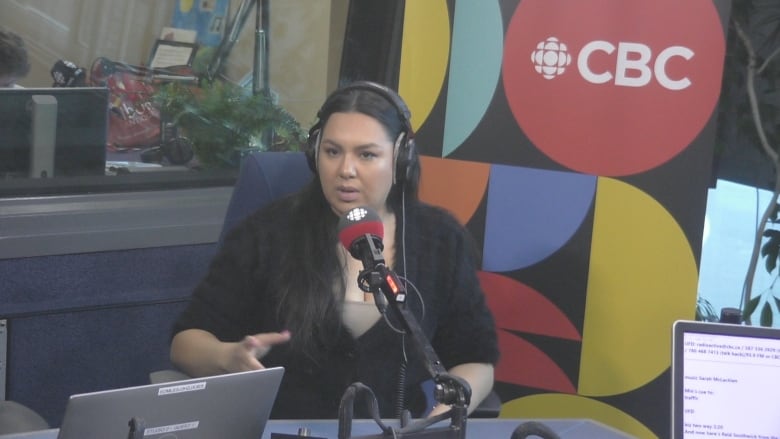 A woman with long black hair and a black blouse sits in front of a microphone in a radio studio. She wears headphones.