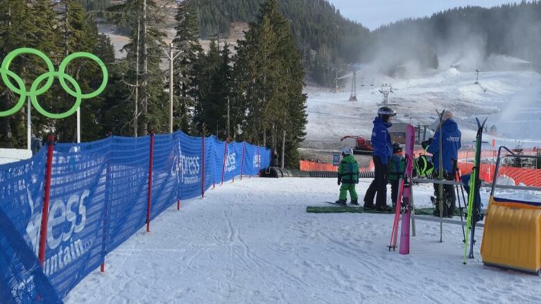 Children and ski instructors in blue uniforms stand at the bottom of a sparsely snow-covered beginners hill.