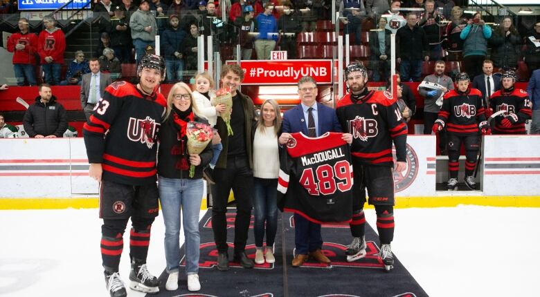 A man in a suit holds up a jersey with 489 written on the back, with sevearal people smiling around him on an ice surface.