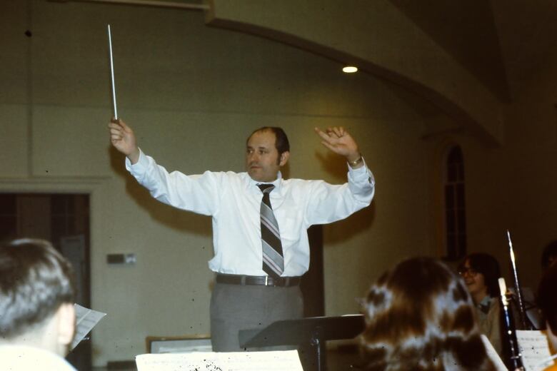 In a vintage photo, a balding man in a white shirt and tie waves a baton in front of several young musicians. 