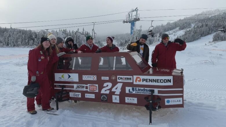 A group of eight people in red overalls stand around a car-shaped toboggan. 