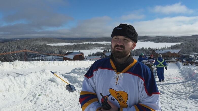 A man in a jersey and a winter hat stands on a ski slope. 