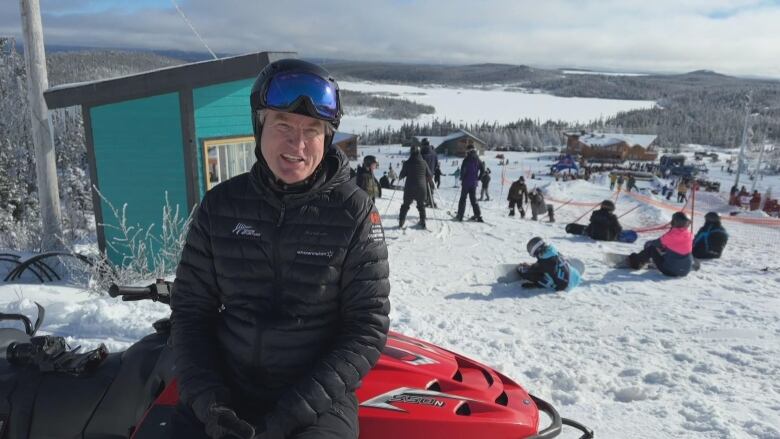 A man in a back jacket and ski goggles sits on a skidoo on a ski slope. 