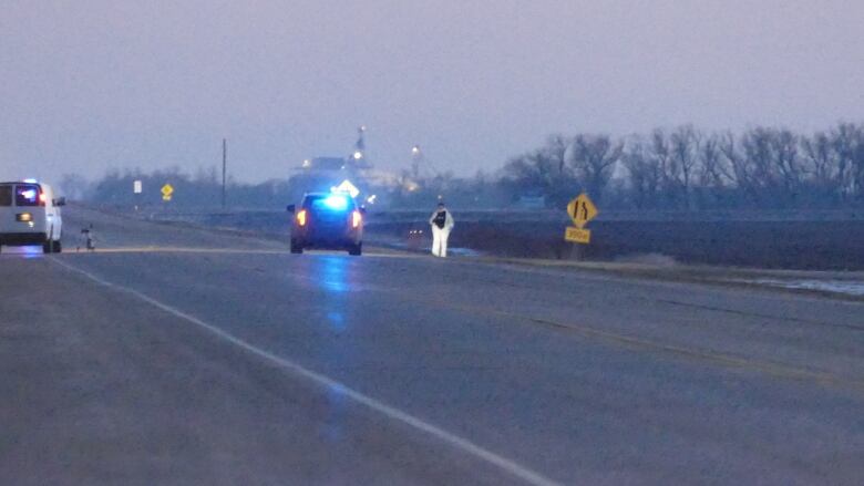 A person walks around in a white coat on a highway near dusk.