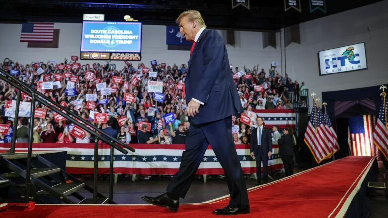 A tall man wearing a dark blue suit, white shirt and red tie, walks toward the left along a red carpet past a crowd of people in bleachers holding signs in the background. 