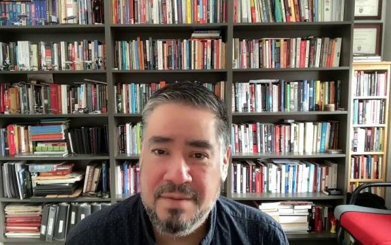 A man looks at a camera with a bookshelf filled with books behind him. He has a salt and pepper beard and a navy blue collared shirt. 