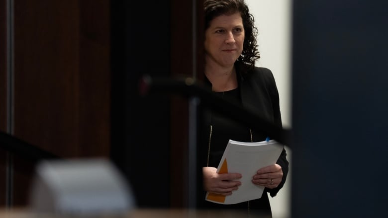 A woman in a dark suit stands in a doorway holding a sheaf of papers.