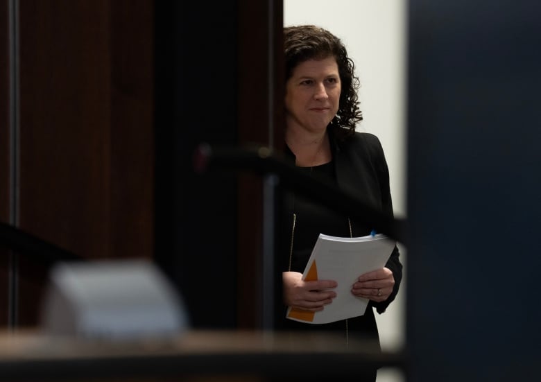 A woman in a dark suit stands in a doorway holding a sheaf of papers.