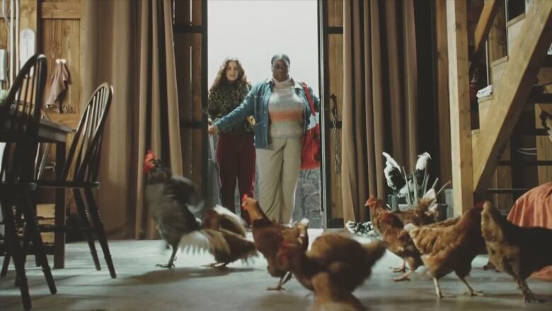 Two women stand in the doorway of a barn that has chickens on the floor.