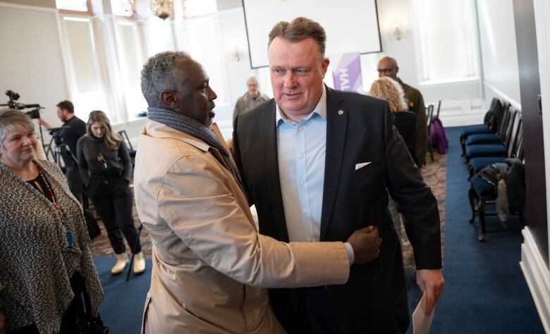 A white man with a suit jacket and white shirt smiles as a Black man in a tan coat hugs him around the waist