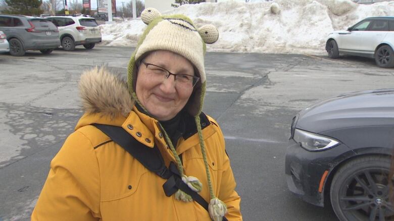 A woman wearing a yellow jacket and frog hat smiles while standing in a parking lot.