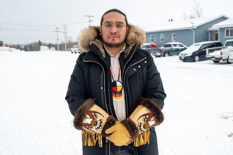 A person wearing a dark winter jacket and moose-hide gloves with a fur trim and beaded eagles on the front of them stands outside on a snowy day.