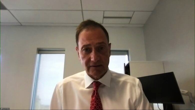 A man in a white collared shirt and a red tie speaks into a web camera