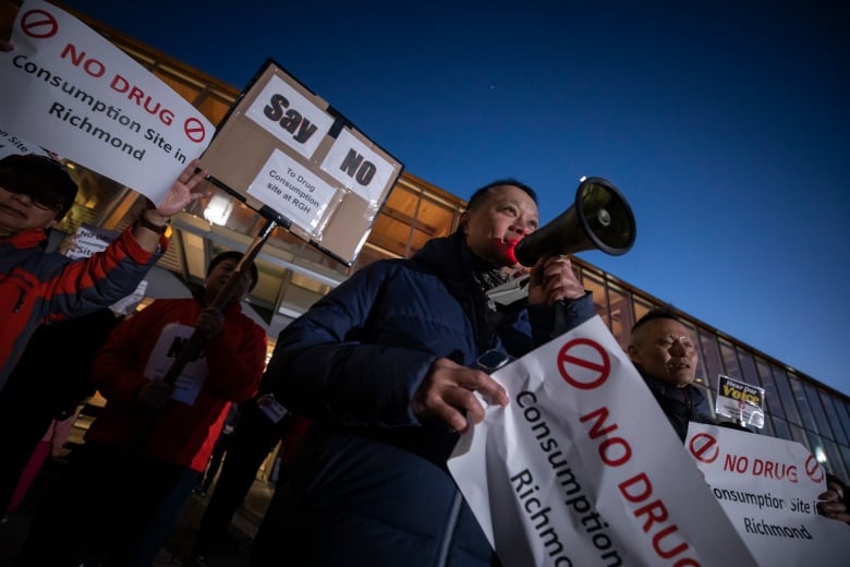 A man holds up a megaphone to his mouth, with people around him holding signs reading 'No Drug Consumption Site in Richmond'.