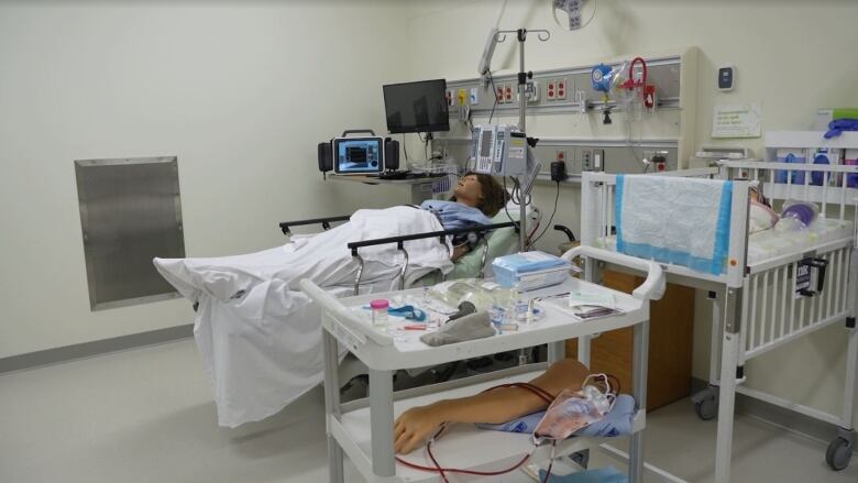 A human-like mannequin with long hair wearing a hospital gown laying in a hospital bed.