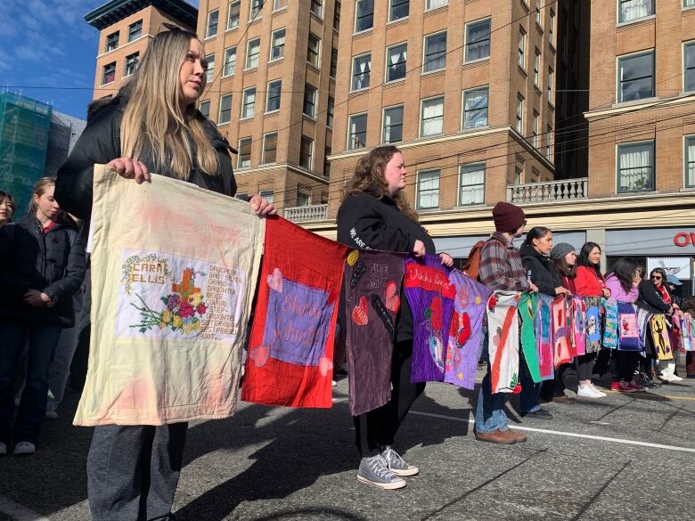 A row of people hold quilted squares with women's names written on them, many with hearts sewn on them, with buildings in the background.