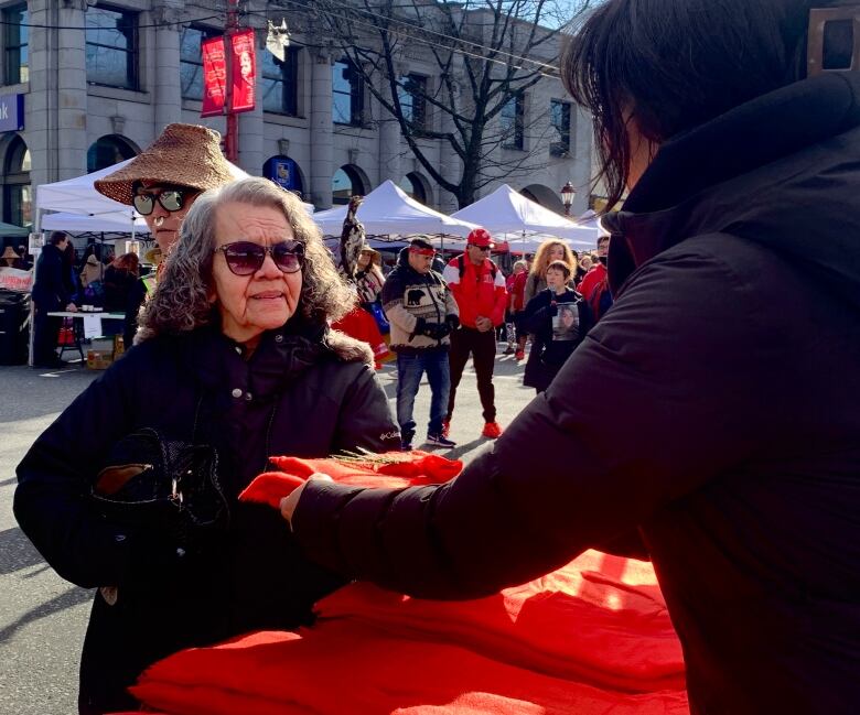 A woman gives another woman a red blanket with a cedar bough on top of it, as a crowd gathers behind them.