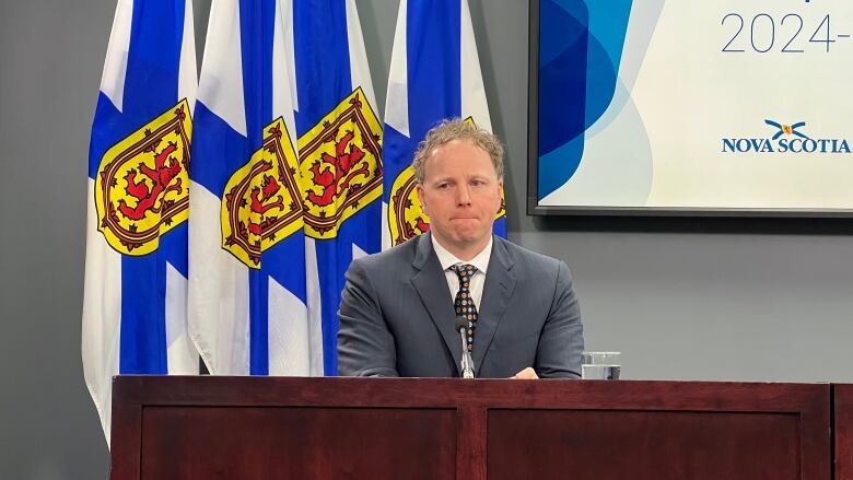  A man in a blue suit sits behind a long podium in front of Nova Scotia flags and a screen with the Nova Scotia government logo on it. 