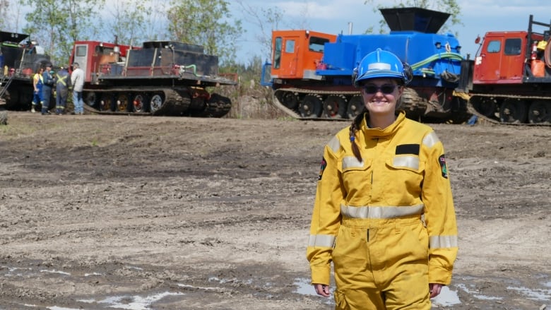 A smiling woman wearing a yellow, long-sleeved protective jumpsuit and a blue hardhat poses in front of heavy machinery in a brown field.