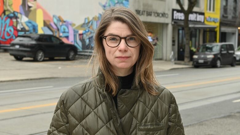 Close-up of woman wearing olive green quilted coat standing on the sidewalk. An empty city street and a row of businesses are behind her. 