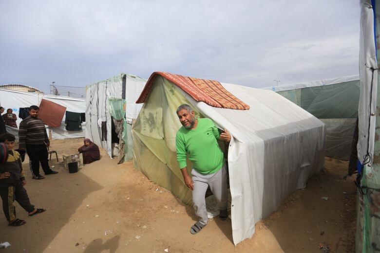 A man in tan pants, grey socks and a green sweater ducks out of the entryway of a tent in an area crowded with other makeshift shelters. 