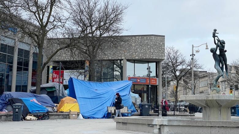 Downtown cement square with a fountain. Several tents and tarps are set up. People are walking through the square. There are no leaves on the trees.