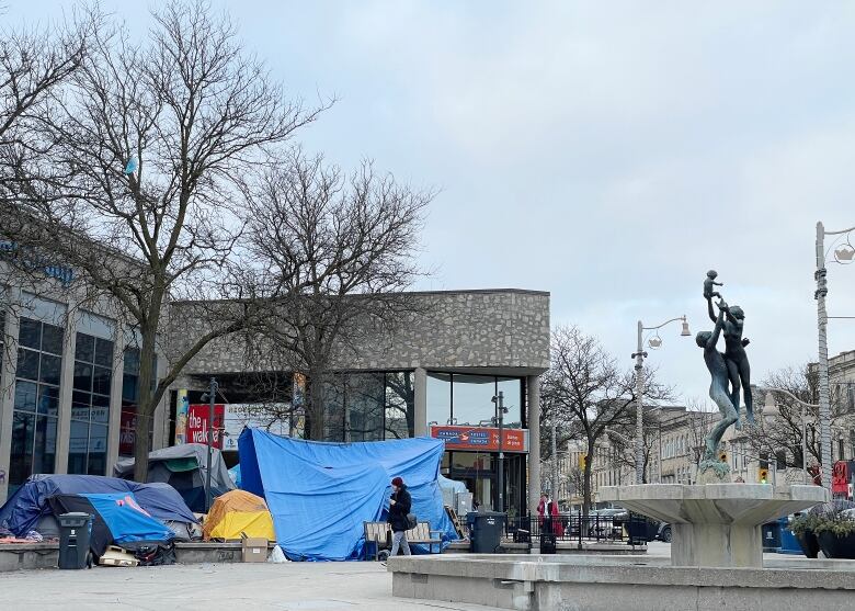 Downtown cement square with a fountain. Several tents and tarps are set up. People are walking through the square. There are no leaves on the trees.