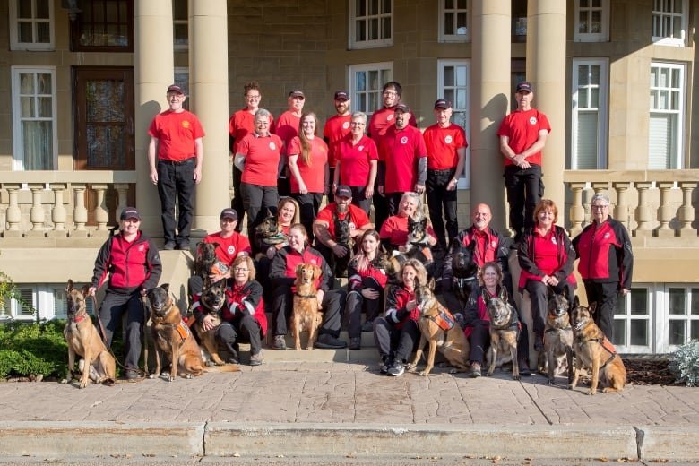 A group of people in red shirts stand with dogs outside a tan building.