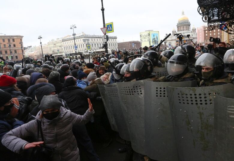 A phalanx of people holding riot shields and wearing riot helmets stand shoulder to shoulder amidst a large crowd in winter clothes, many with their faces covered.