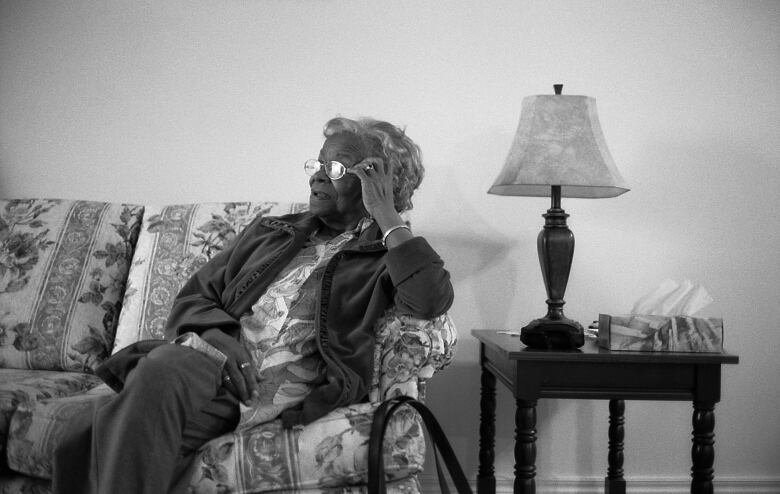 A black-and-white photo of an elderly woman sitting on a couch, looking away from the camera.