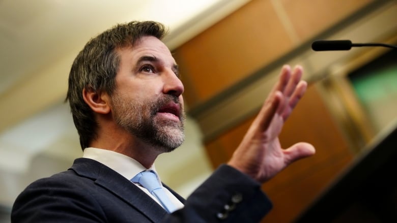 A man wearing a suit with a beard talks at a podium.