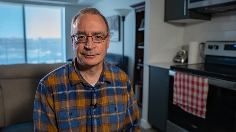 A man with glasses and a plaid shirt sits and apartment with a window in the background and a stove on the right.