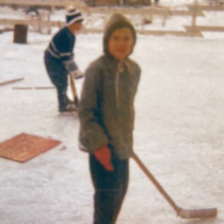 Old photograph shows two children on ice. Child in foreground is looking toward the camera, smiling, while holding a hockey stick.