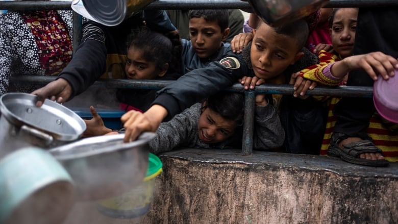 Children hold out bowls for food from behind a barrier.