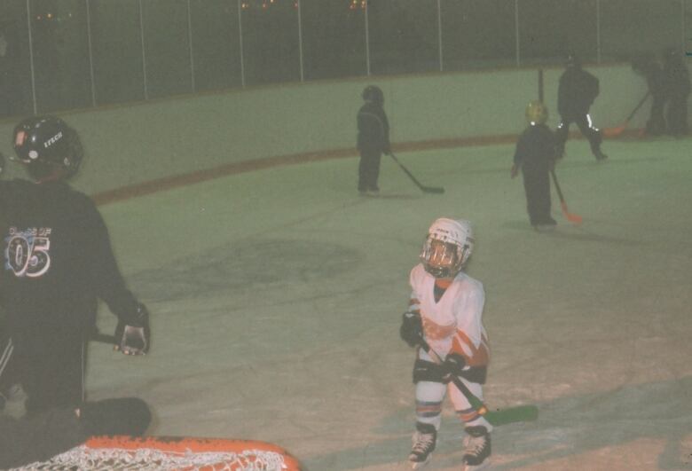 A child wears hockey gear and a helmet carrying a stick on an ice rink with other children nearby with sticks and gear.