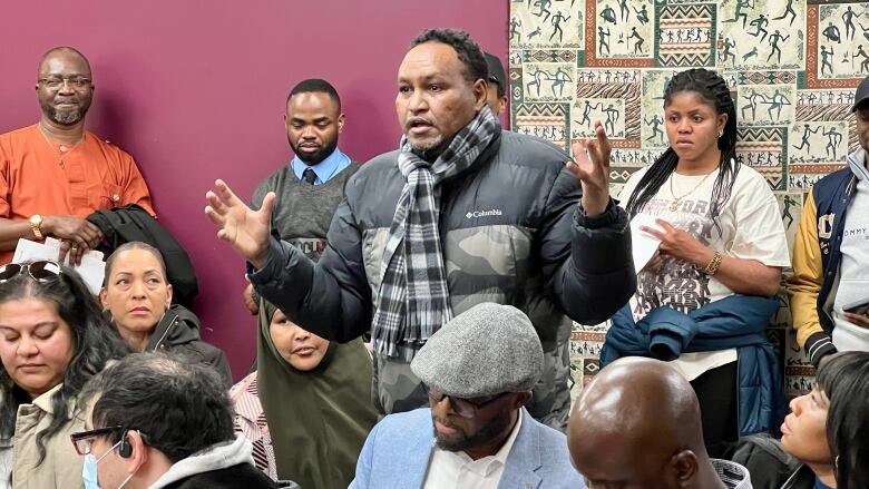 A man gesturing while talking. He's surrounded by a big group of people, some sitting, some standing.