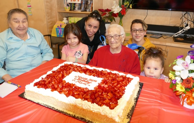 An elderly woman smiles at the camera while sitting in front of a big birthday cake and surrounded by three adults and two children.