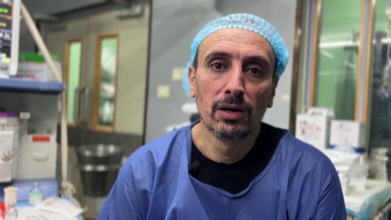 A man in a blue hospital smock and matching hair cap looks directly into the camera.