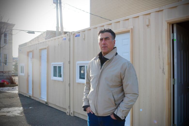  A man in a beige jacket stands in front of a row of beige shipping containers