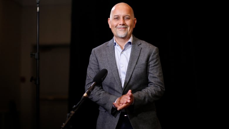 A bald Indigenous man with a white goatee wearing a grey jacket smiles while standing in front of a mic.